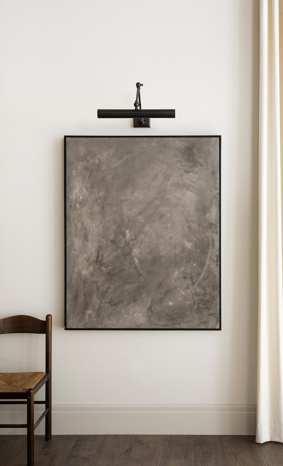 A large, abstract gray painting with subtle texture hangs on a white wall under a black picture light. A wooden chair with a woven seat is partially visible to the left, and a cream curtain is to the right, by McGee & Co.