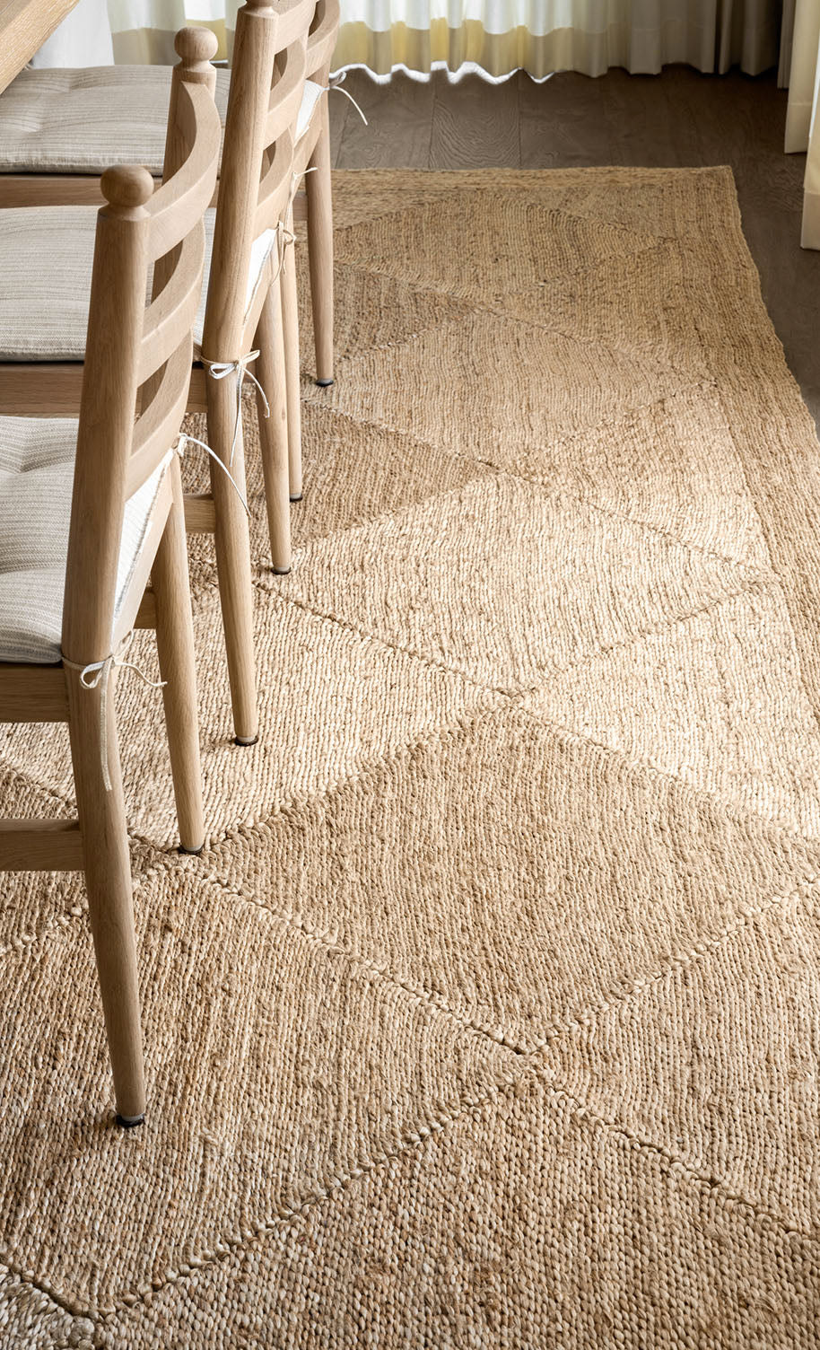 A close-up view of a textured, woven beige rug beneath wooden chairs with light-colored seat cushions, placed next to a table in a sunlit room by McGee & Co.