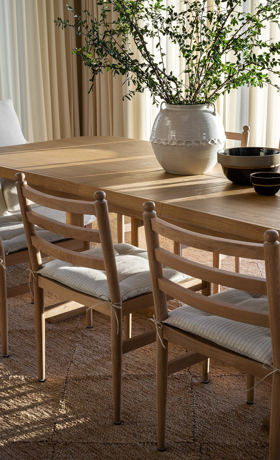 A sunlit wooden dining table with matching chairs, a white ceramic vase holding green branches, and black bowls on a woven rug, with light curtains in the background, by McGee & Co.