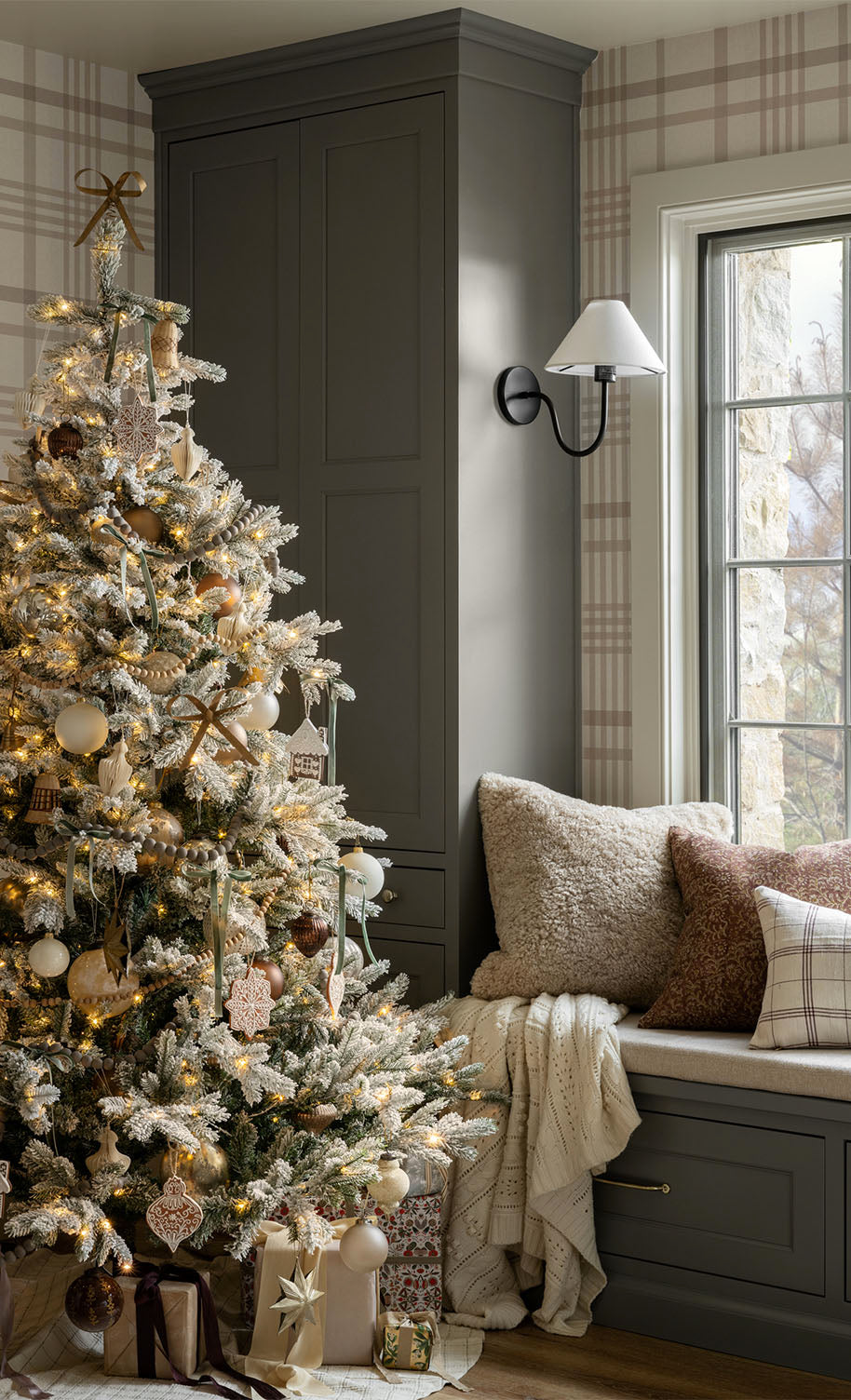 A decorated Christmas tree with lights and ornaments stands by a window seat with cozy pillows and blankets; wrapped gifts are placed under the tree in a softly lit, plaid-patterned room, by McGee & Co.