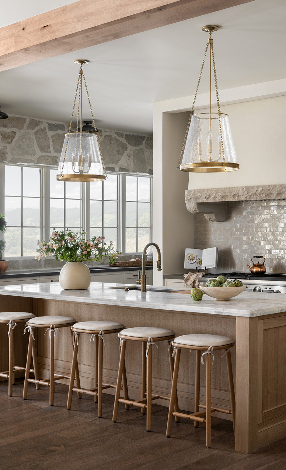 A bright, modern kitchen with light wood cabinets, a marble island with four wooden stools, pendant lights, a stone backsplash, large windows, and a vase of flowers on the counter by McGee & Co.