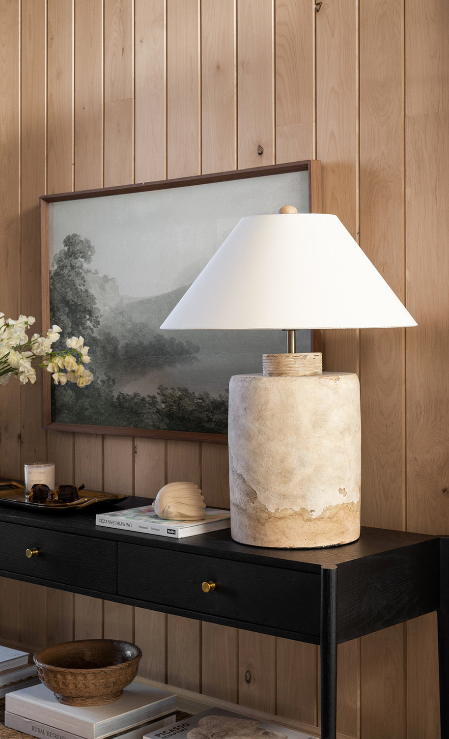 A rustic ceramic lamp with a white shade sits on a black console table, beside stacked books and a white flower arrangement, against a wood-paneled wall with a framed landscape painting, by McGee & Co.