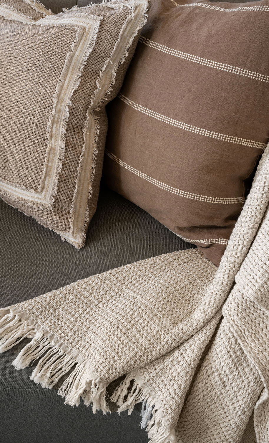Two textured throw pillows, one with fringe and one with white stripes, rest on a dark sofa alongside a chunky woven cream-colored throw blanket with fringed edges, by McGee & Co.