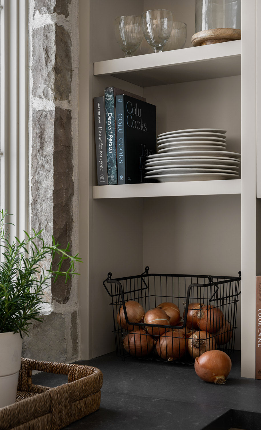 Open shelving in a kitchen with stacked white plates, cookbooks, glassware, a wire basket of onions, a potted plant, and a stone wall beside a window by McGee & Co.