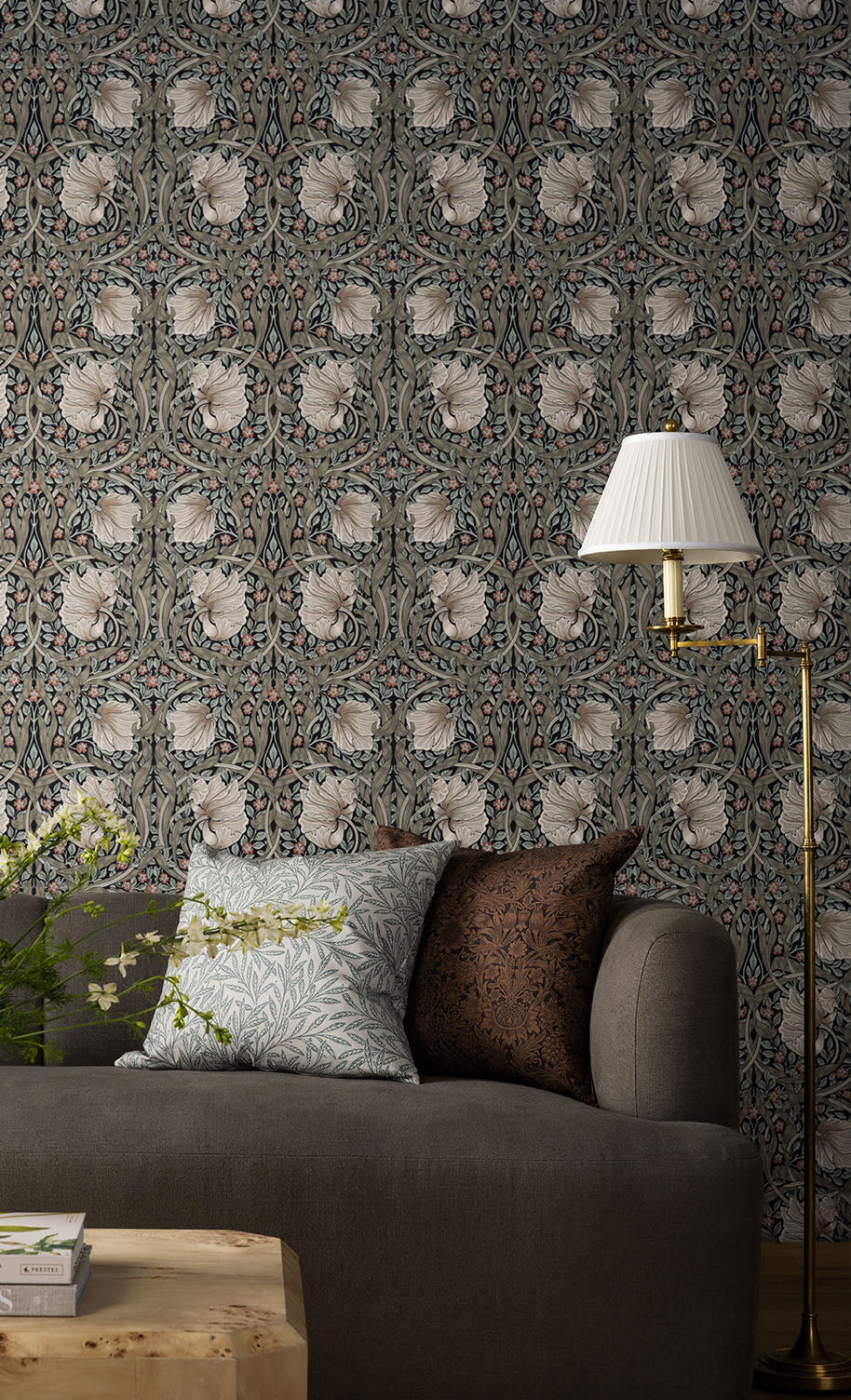 A gray sofa with patterned cushions sits in front of a detailed floral wallpaper, next to a wooden table and a gold floor lamp with a white pleated lampshade, by McGee & Co.