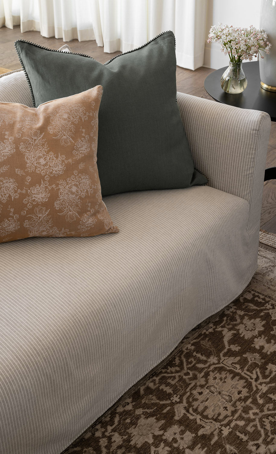 A light gray pinstriped sofa with a green cushion and a beige floral-patterned cushion, next to a round black side table with a vase of flowers, on a patterned rug by McGee & Co.