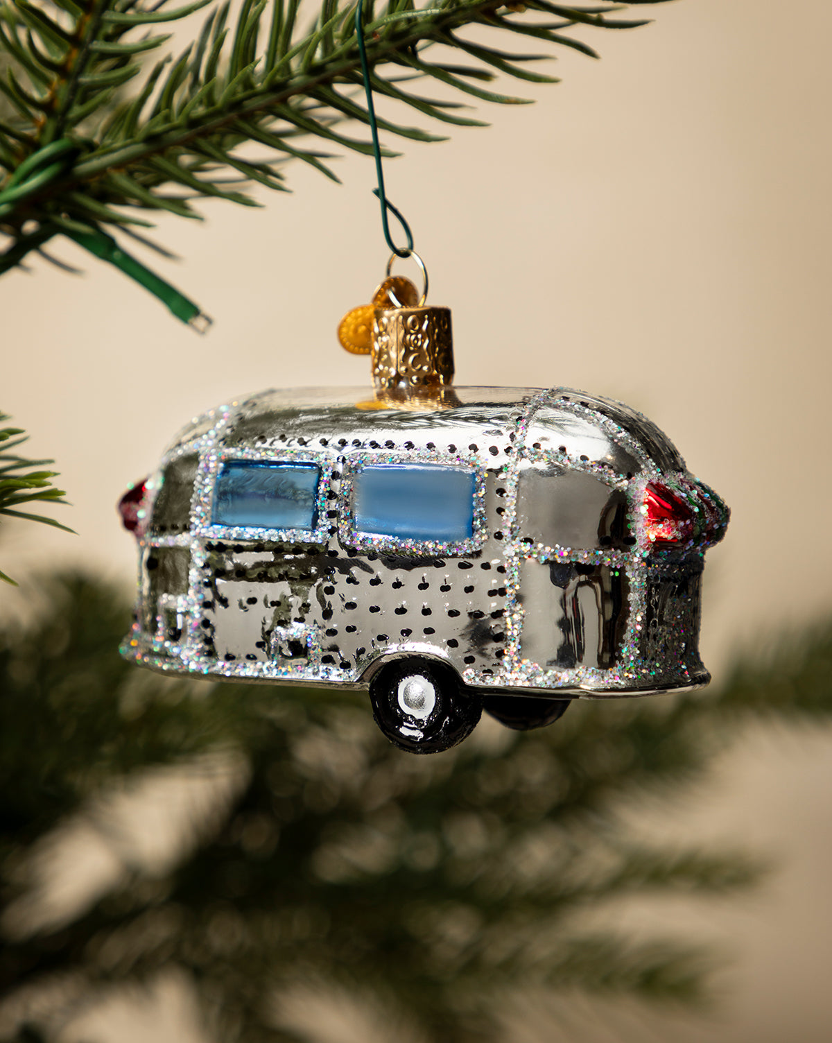 The Vintage Trailer Ornament features shiny silver paint, blue windows, and glitter accents, bringing retro holiday charm as it hangs from a Christmas tree branch, by McGee & Co.