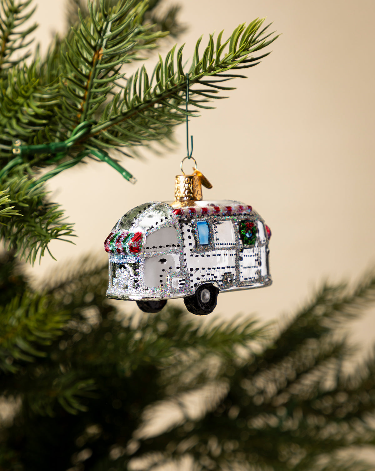 The Vintage Trailer Ornament, designed as a silver camper with festive red and green accents, hangs on a green pine tree branch against a neutral background, by McGee & Co.