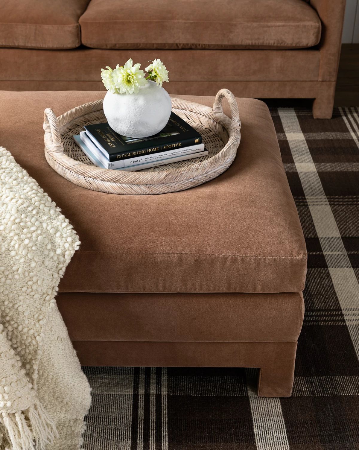 The Makerspalm Appoline Ottoman in tan is topped with a woven tray holding a white vase of flowers and two books, set next to a textured cream blanket on a brown and cream plaid rug, McGee & Co.
