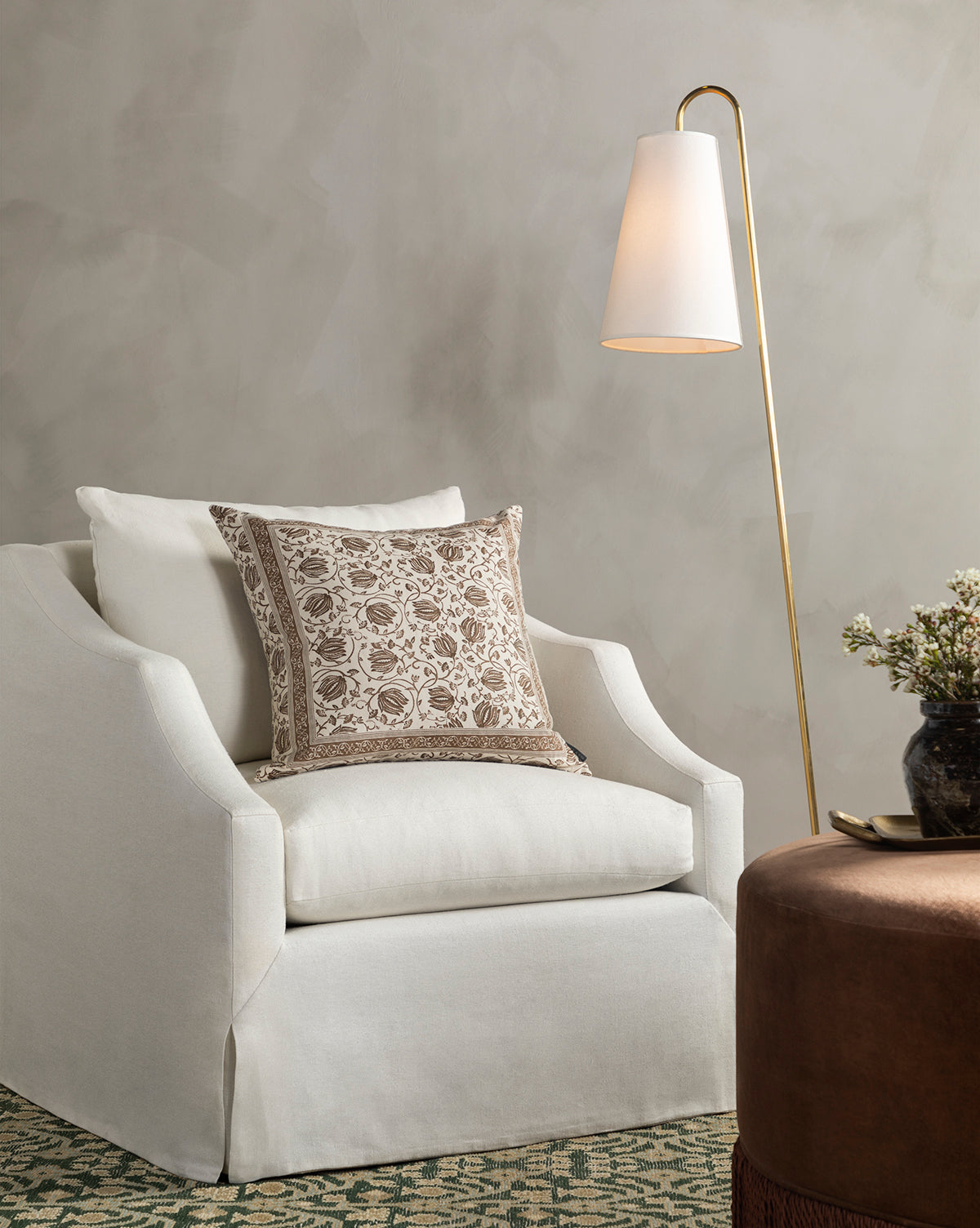 A cozy Everleigh Slipcover Lounge Chair by Community with a patterned beige cushion sits beside a gold floor lamp with a white shade, while a round brown ottoman and floral vase complete the elegant, neutral-toned decor. McGee & Co.
