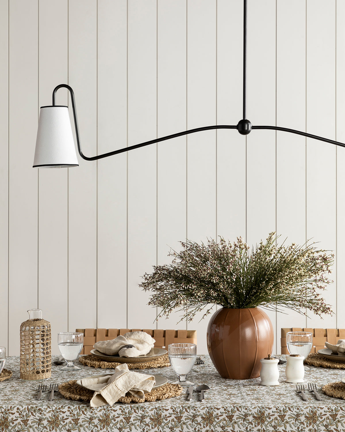 A dining table set with plates, glasses, and woven placemats sits beneath a modern black pendant light. At the center is the Idris Vase filled with flowers, placed on a floral-patterned tablecloth.