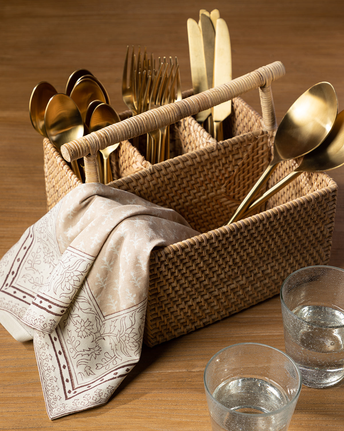 A Rattan Utensil Caddy filled with gold-colored cutlery, a patterned beige napkin over its side, and two glasses of water are arranged on a wooden table by McGee & Co.