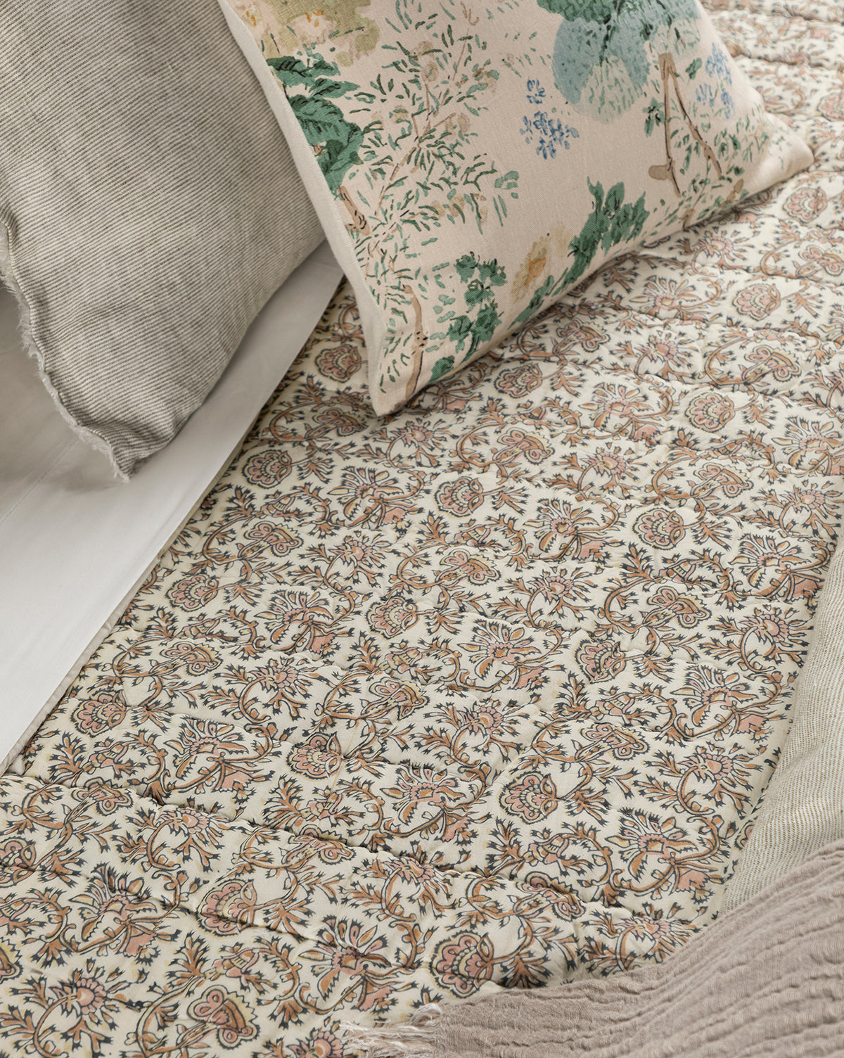 A close-up of a bed with the Leander Quilt, showcasing intricate floral and paisley patterns, is paired with beige and green botanical-print pillows to create an inviting bedding arrangement.