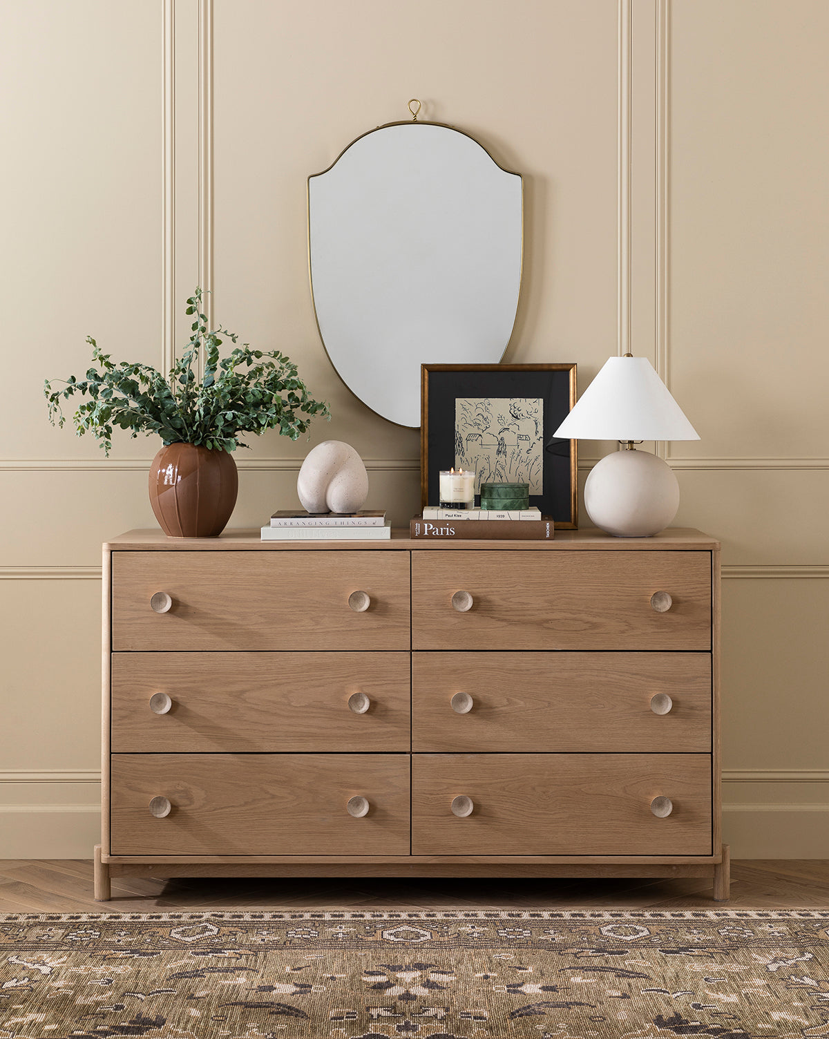 A light wood dresser with six drawers stands by a beige paneled wall. On top are an Idris Vase with greenery, decorative objects, framed art, books, a lit candle, and a white lamp. Above hangs a shield-shaped mirror.