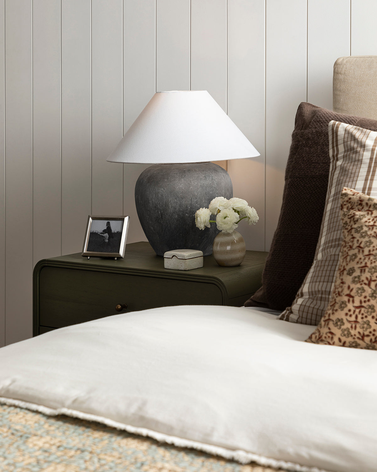 A cozy bedroom nightstand with the Montague Table Lamp in antique black, a small vase of white flowers, a framed photo, a decorative box, and neatly arranged neutral-toned pillows and bedding by McGee & Co.