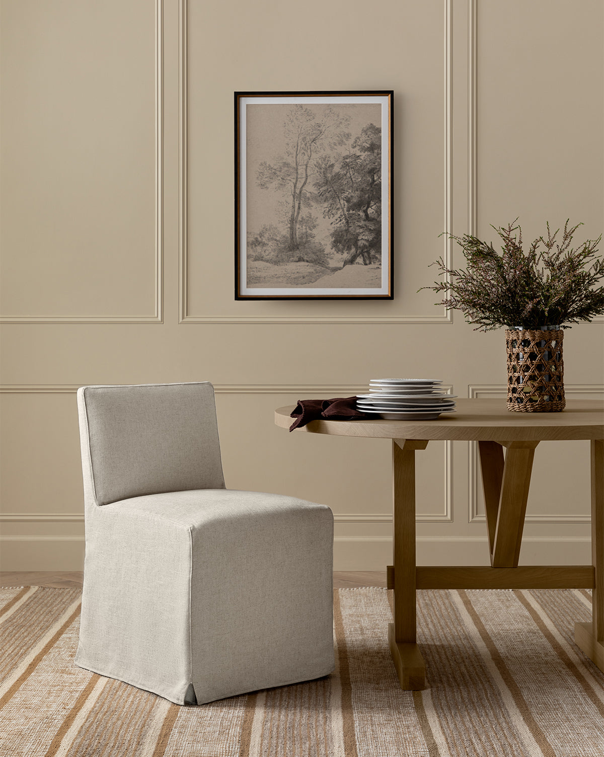 A minimalist room features the Olivier Slipcover Dining Chair, a wooden table with stacked plates and a vase of greenery, a striped rug, paneled walls, and a framed landscape artwork above, by McGee & Co.