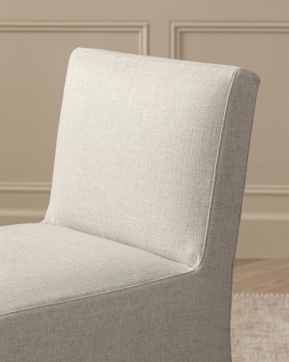 A close-up of the Olivier Slipcover Dining Chair in light beige fabric, with a feather and down cushion, set against a beige wall with decorative molding and part of a woven rug visible on the floor, by McGee & Co.
