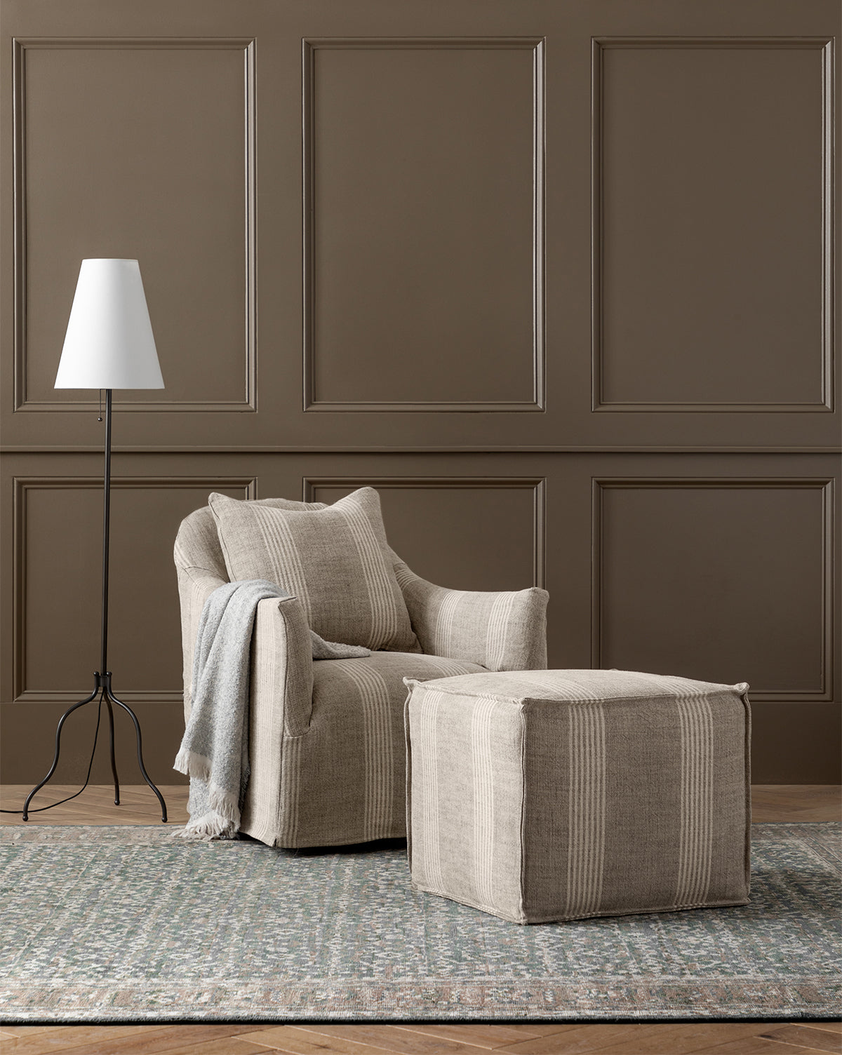 A cozy beige armchair from the made-to-order Johannes Cube collection, with a cushioned seat and matching ottoman, sits on a patterned rug by a modern white floor lamp against a brown paneled wall. A folded gray throw rests on the chair’s arm, by McGee & Co.