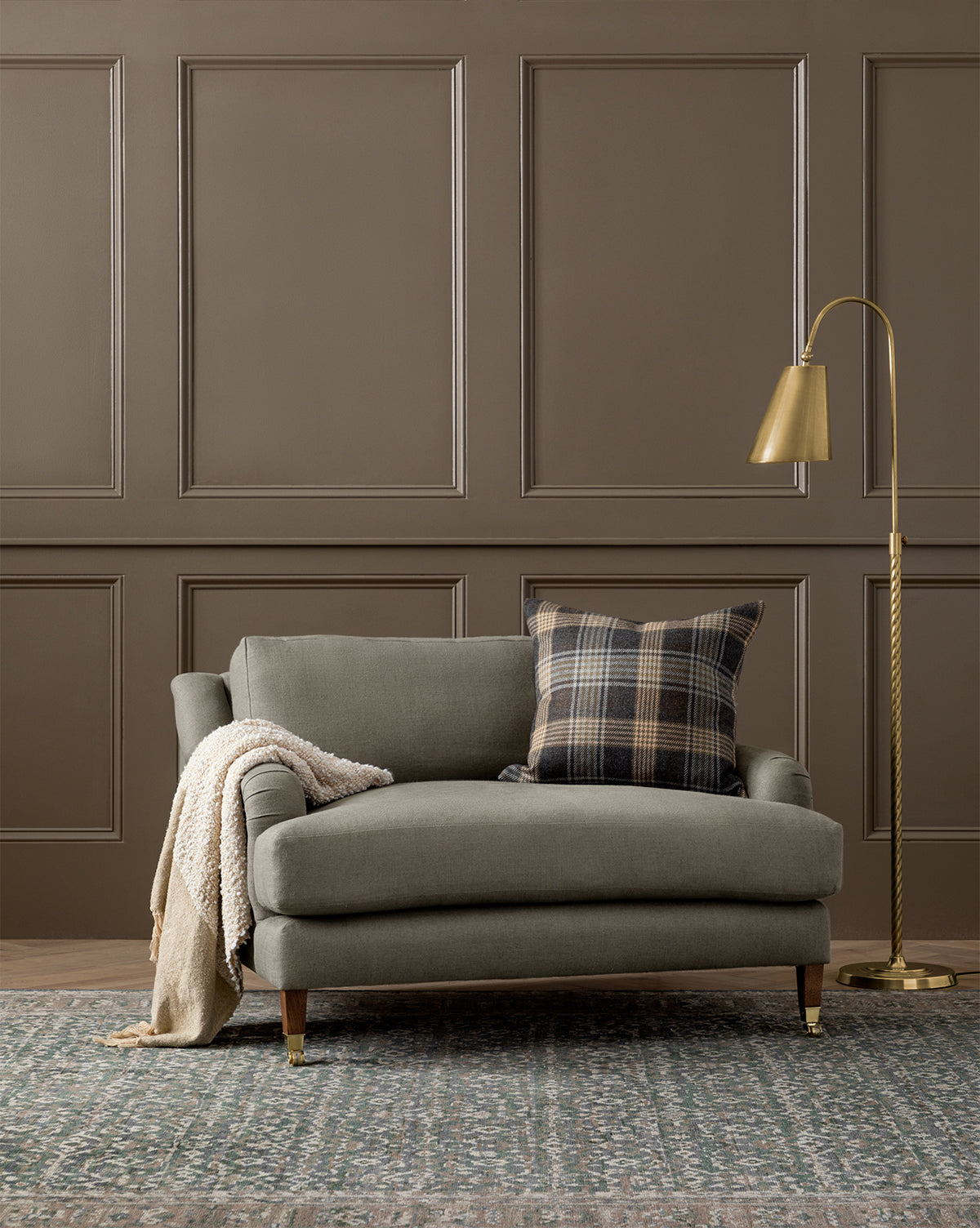 The Community Lucille English Roll Arm Chair and a Half features plush down-wrapped cushions. Shown in cozy gray with a plaid pillow and cream knit blanket, its set on a patterned rug beside a gold floor lamp and brown paneled wall. McGee & Co.