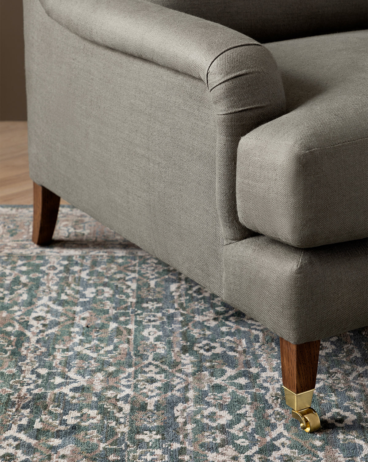 Close-up of a gray upholstered sofa with wooden legs and brass casters, beside the Lucille English Roll Arm Chair and a Half with a plush down-wrapped cushion, all set on a blue, beige, and green patterned area rug, by McGee & Co.