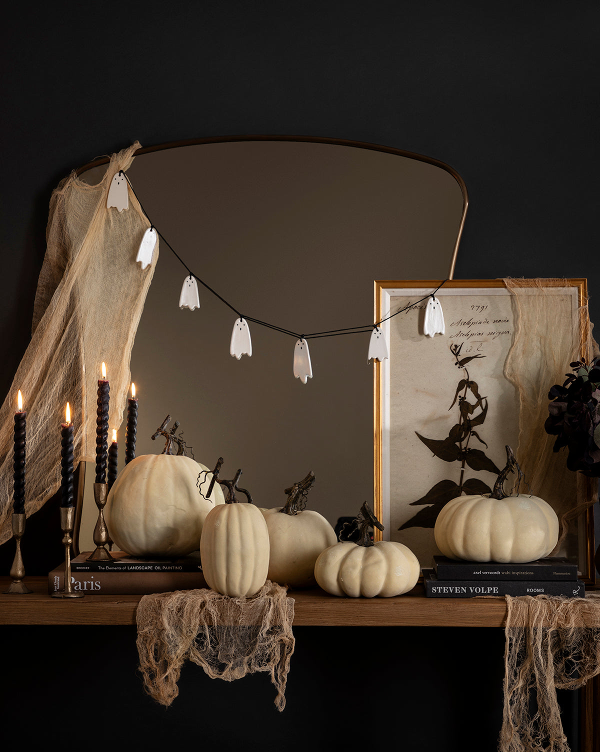 A Halloween mantel features a 3' Ceramic Ghost Garland, white pumpkins, black candles, ghost string lights, draped gauze, vintage art leaning against a mirror—all set against a dark backdrop, by McGee & Co.