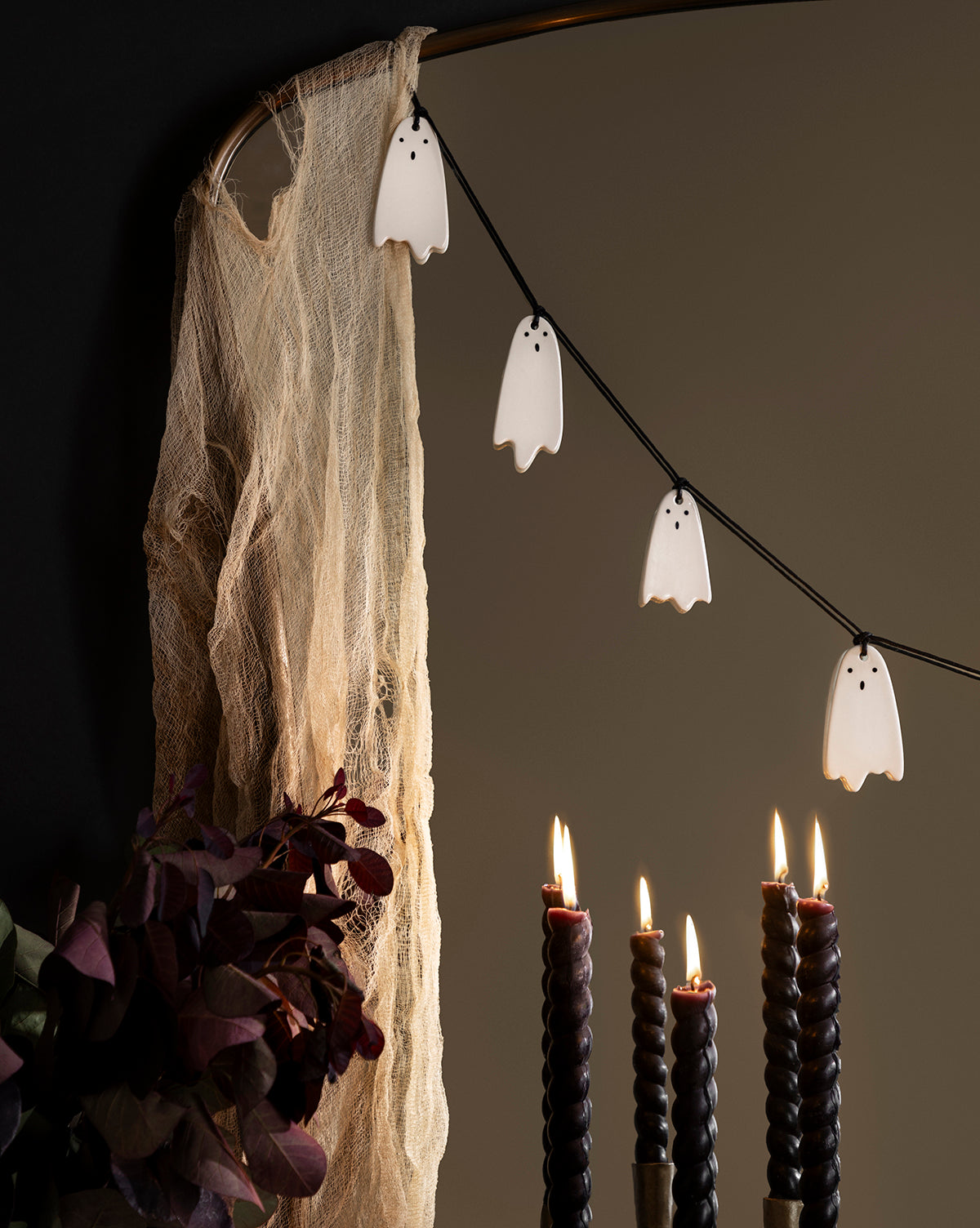 A 3' Ceramic Ghost Garland hangs beside tattered beige fabric on a dark wall. In front, five black twisted candles burn as dark red foliage adds a finishing touch to the Halloween decor, by McGee & Co.