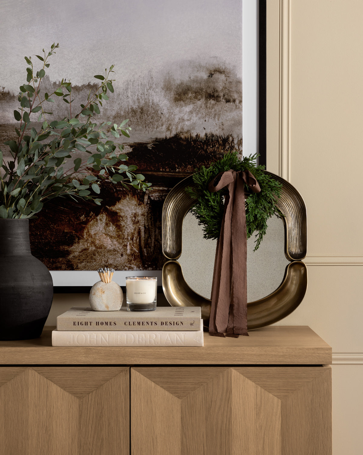 A wooden sideboard displays a black vase with green foliage, stacked books, a candle, matches, and a cast aluminum sculpture with a wreath and ribbon, set against the Salvatore Wall Mirror, by McGee & Co.