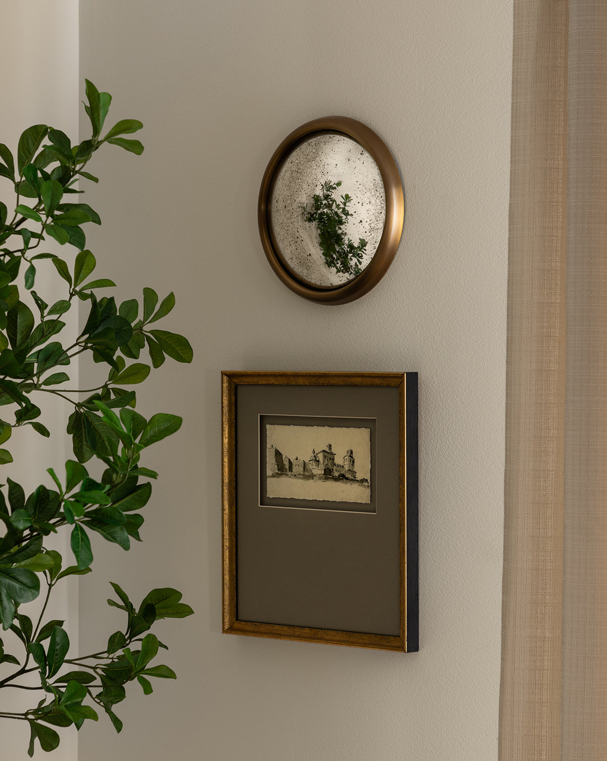 A leafy plant rests by a white wall with two framed pieces: a round gold-framed mirror above gallery-quality artwork titled Sepia City, both hanging in the corner near a beige curtain.