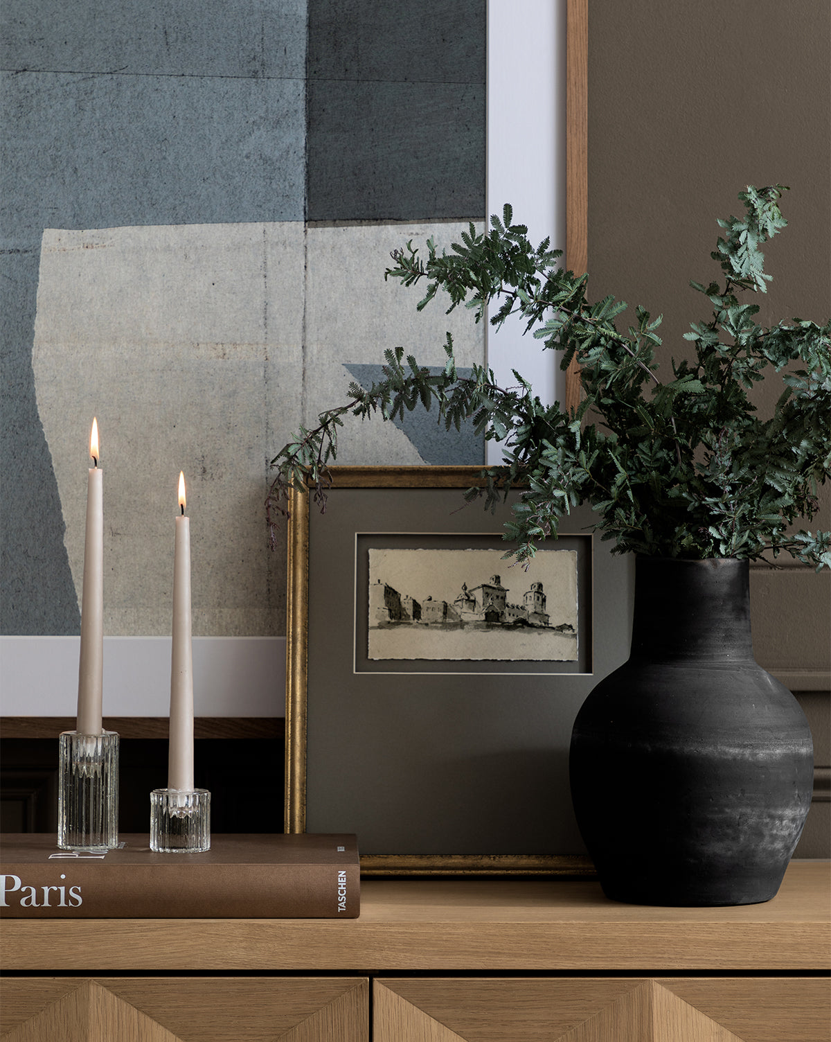 A modern wooden sideboard displays two lit candles in glass holders, a brown Paris book, a black vase with greenery, and the Sepia City gallery-quality artwork featuring a romantic town scene against a neutral-toned wall.