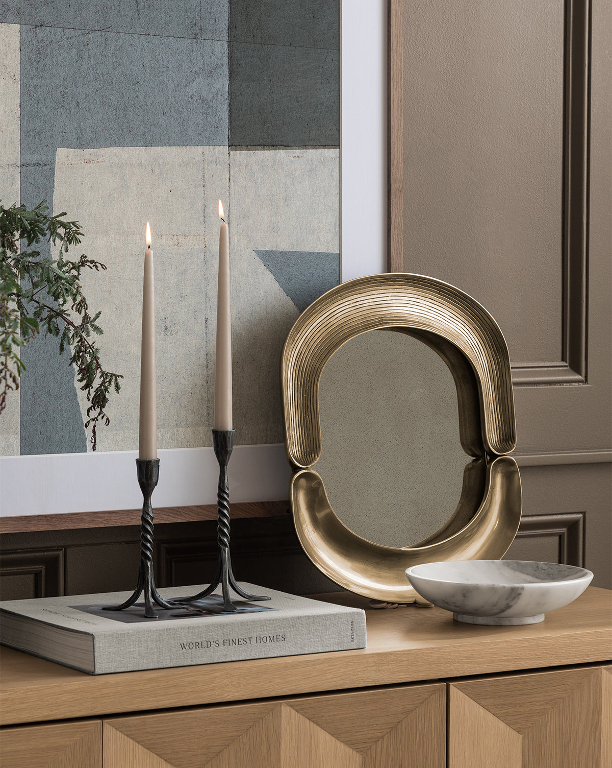 A modern still life with the Salvatore Wall Mirror, two lit candles in black holders on a book, a wavy tray in antique gold finish, a white marble bowl, and abstract art atop a wooden cabinet against a paneled wall, by McGee & Co.