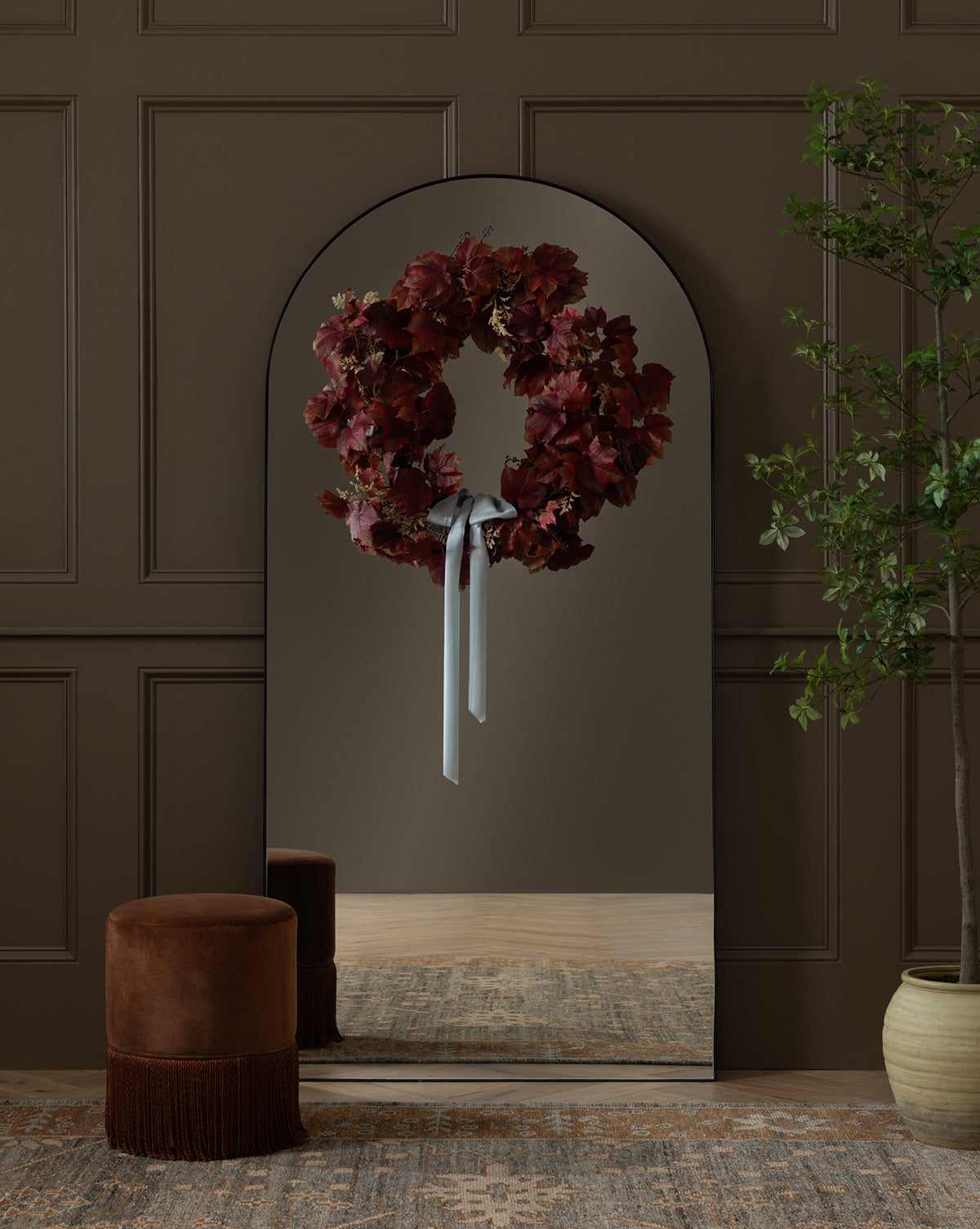 A tall, arched mirror reflects the Faux Autumn Grapevine Wreath accented with deep red flowers and a white ribbon. In front are a brown velvet stool and patterned rug, plus faux greenery to the right for seasonal decor.