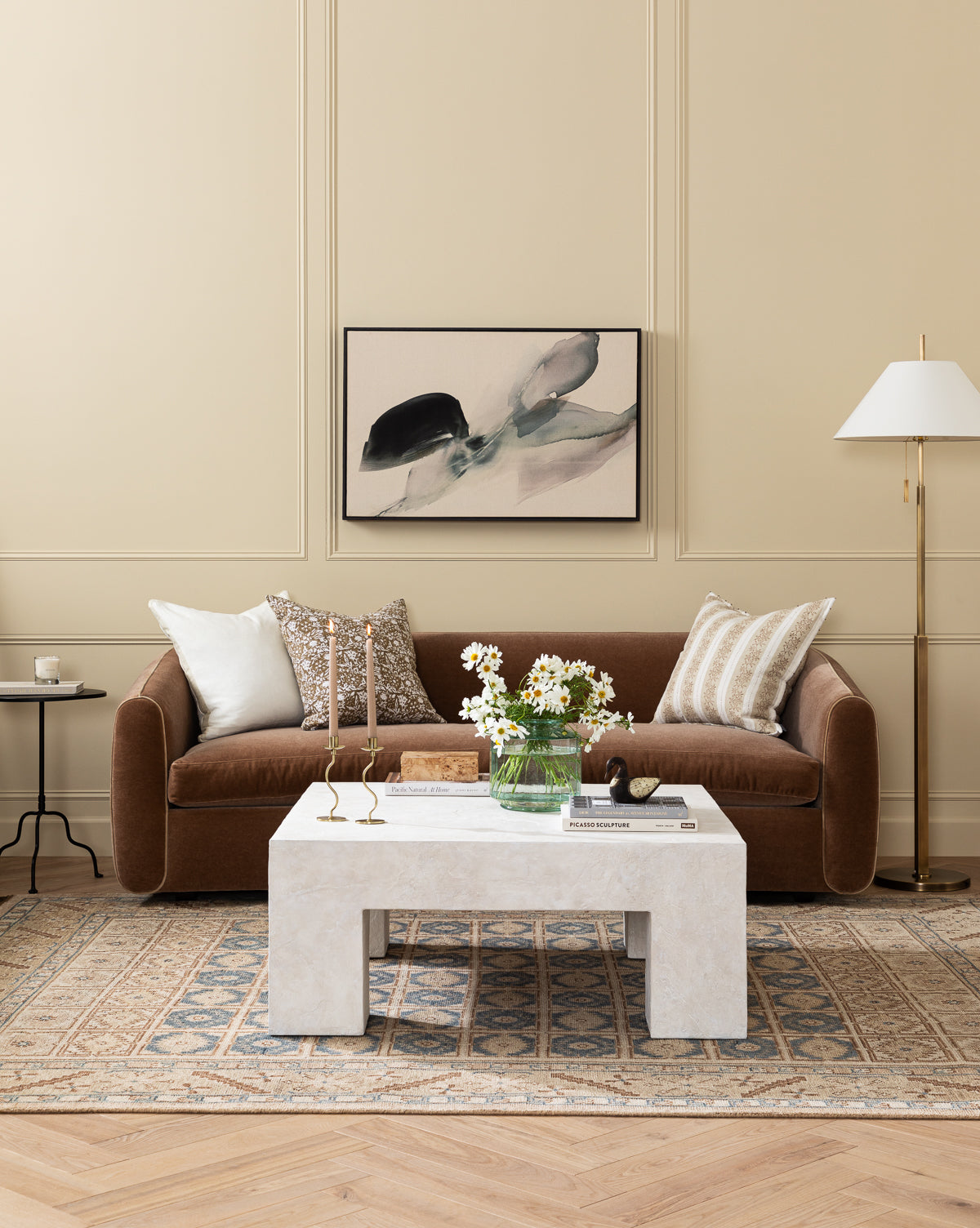 A stylish living room with a brown velvet sofa, neutral pillows, a marble coffee table with flowers, gold candlesticks, an abstract painting above the sofa, a modern floor lamp, and the Nunez Hand-Knotted Wool Rug on a wooden floor, by McGee & Co.