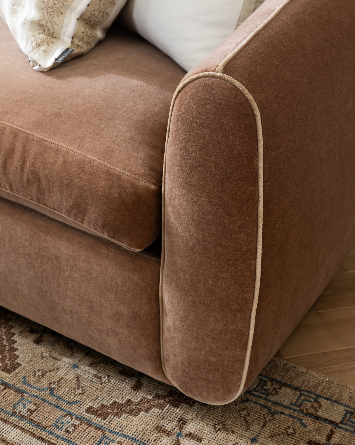 A close-up of the Wexler Sofa 90 in customizable designer fabric, shown with brown upholstery and light piping on the armrest, set on a patterned rug with cushions partially visible in the background, by McGee & Co.