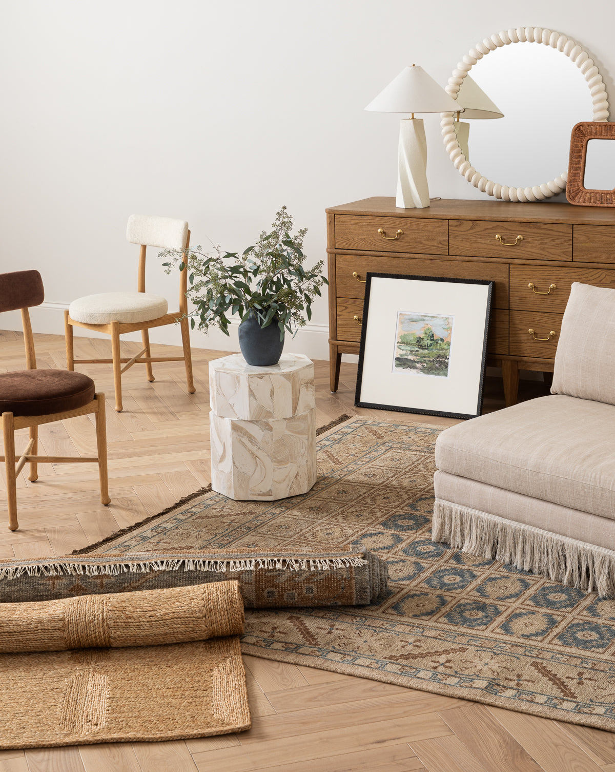 A cozy living room features a wooden dresser with a round mirror, two chairs, a beige fringed lounge seat, the Nunez Hand-Knotted Wool Rug, a small white side table with a plant, and framed artwork leaning against the wall, by McGee & Co.