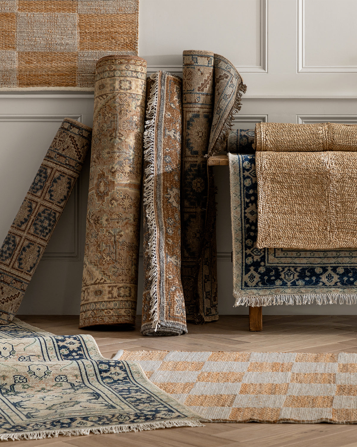 Tulie Hand-Knotted Wool Rugs in neutral hues, some rolled, others folded or laid flat, are displayed against a pale paneled wall and wooden floor, by McGee & Co.