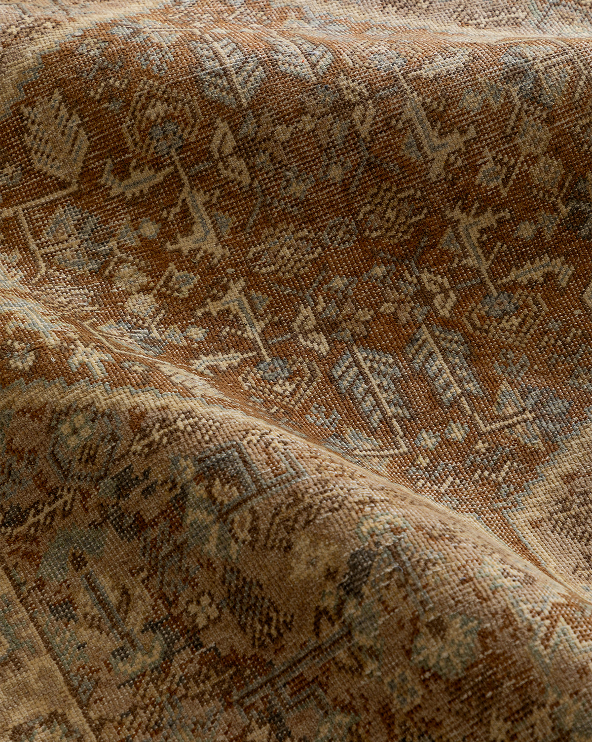 A close-up of the Tulie Hand-Knotted Wool Rug displays its textured, brown-toned wool with intricate geometric and tribal designs, featuring gentle folds and color variations from tan to muted blue accents, by McGee & Co.