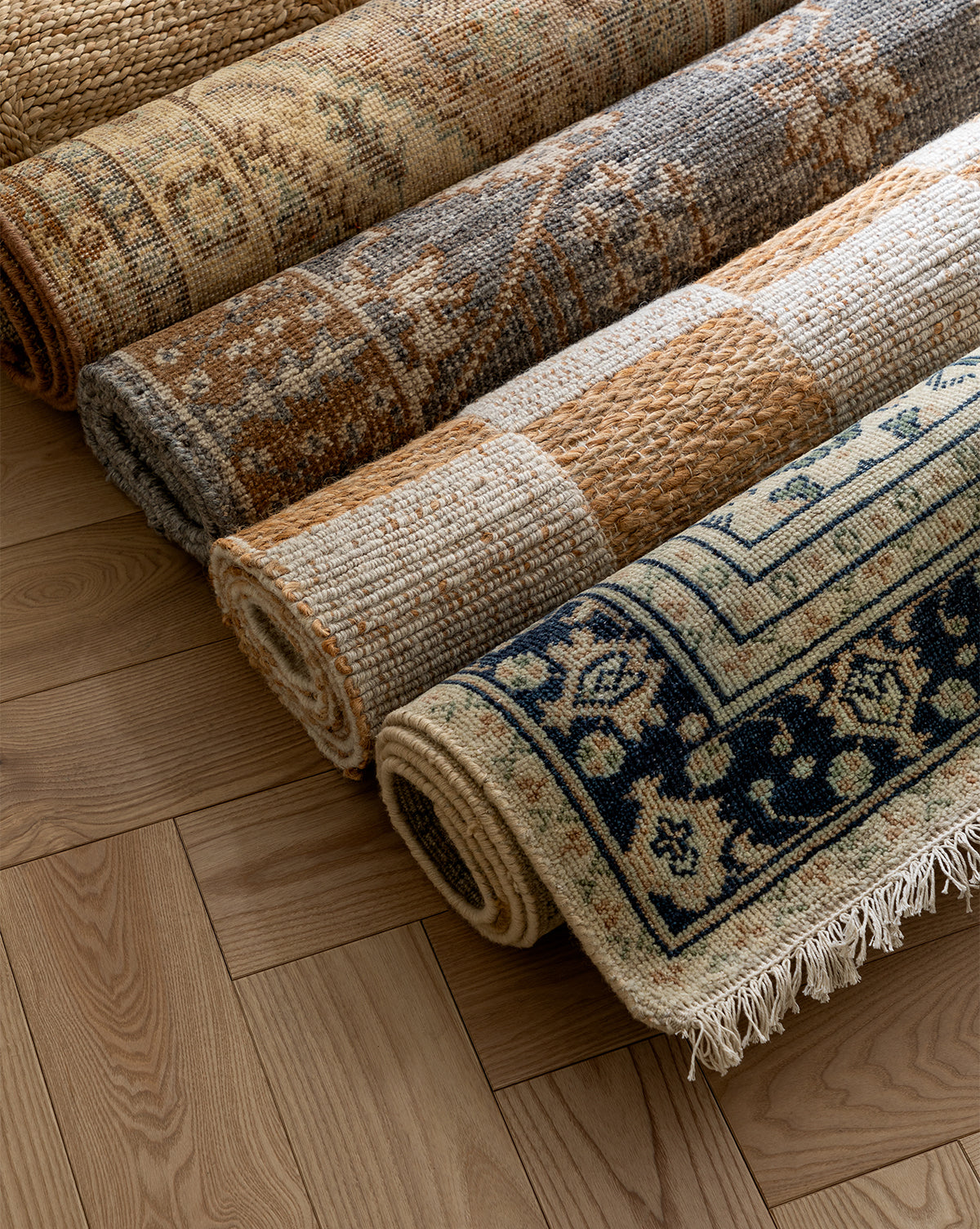 Four rolled-up rugs, including the Vercelli Hand-Knotted Wool Rug, rest on a light herringbone floor. The wool rugs display geometric, floral, and checkered patterns in earthy beige, blue, and brown hues, by McGee & Co.