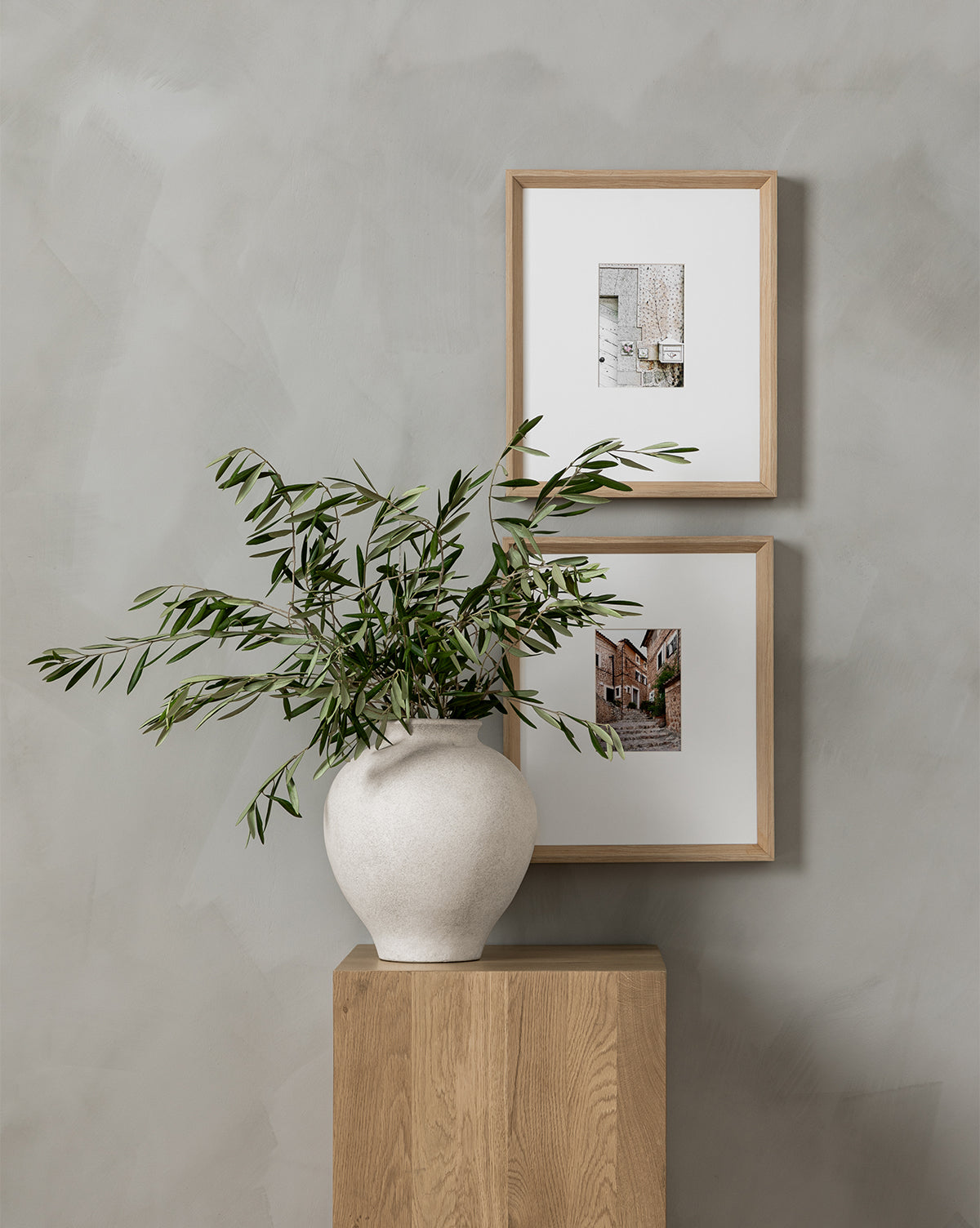 A Krissan Vase with a pebbled white finish displays green branches on a wooden pedestal, while two framed building photos hang on the grey textured wall behind, by McGee & Co.