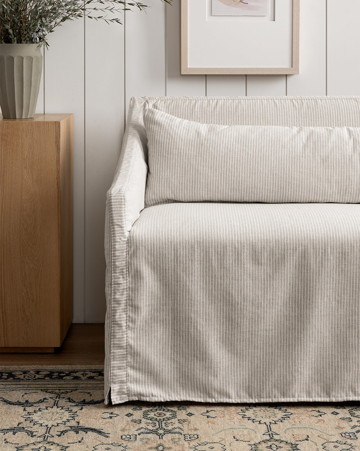 An Olivier Slipcover Dining Bench with light-colored stripes and feather-and-down cushions sits by a wooden side table with a potted plant. Above, a framed picture hangs on a white paneled wall over a patterned rug, by McGee & Co.