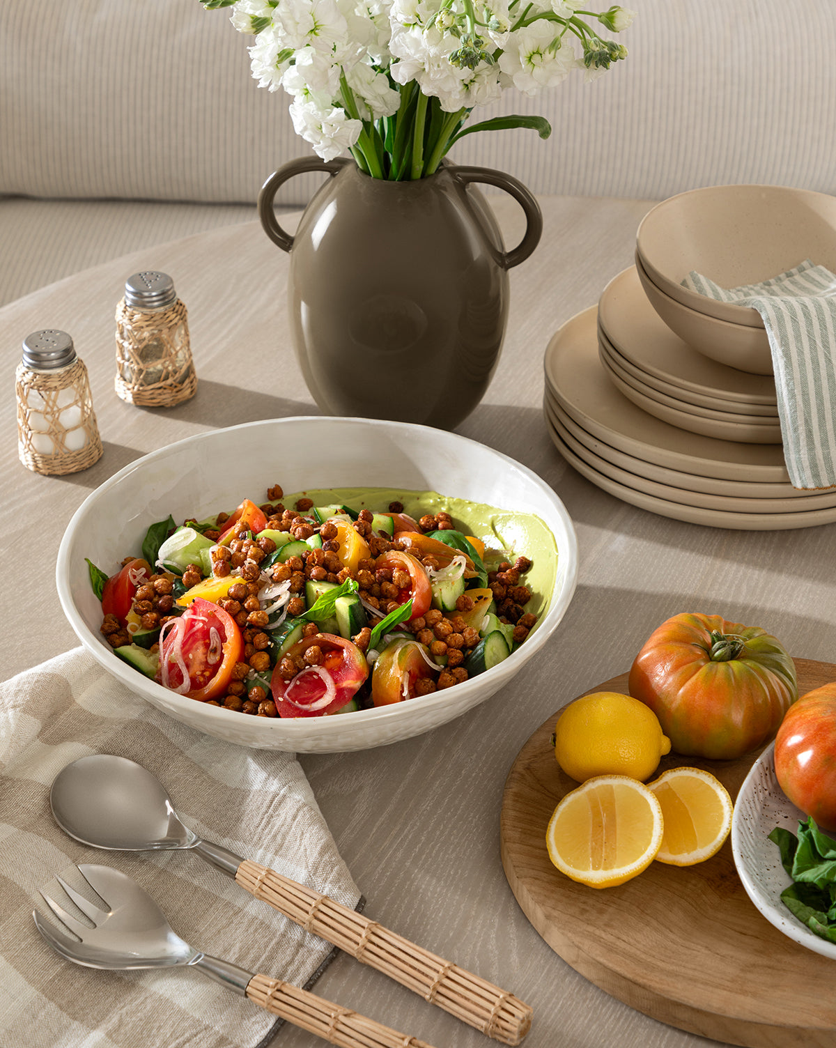 A fresh salad in a white bowl sits on a table with lemons, heirloom tomatoes, plates, utensils, salt and pepper shakers, and the Leylani Vase filled with white flowers.