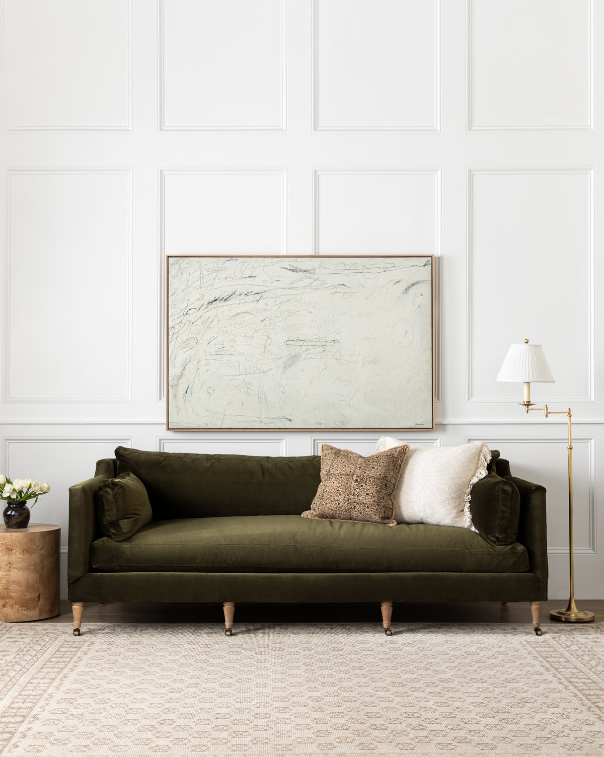 A green velvet sofa with decorative cushions faces a white paneled wall and an abstract painting, flanked by a side table with flowers and a floor lamp, all anchored by the Anya Neutral Hand-Knotted Wool Rug in a subtle beige pattern, by McGee & Co.
