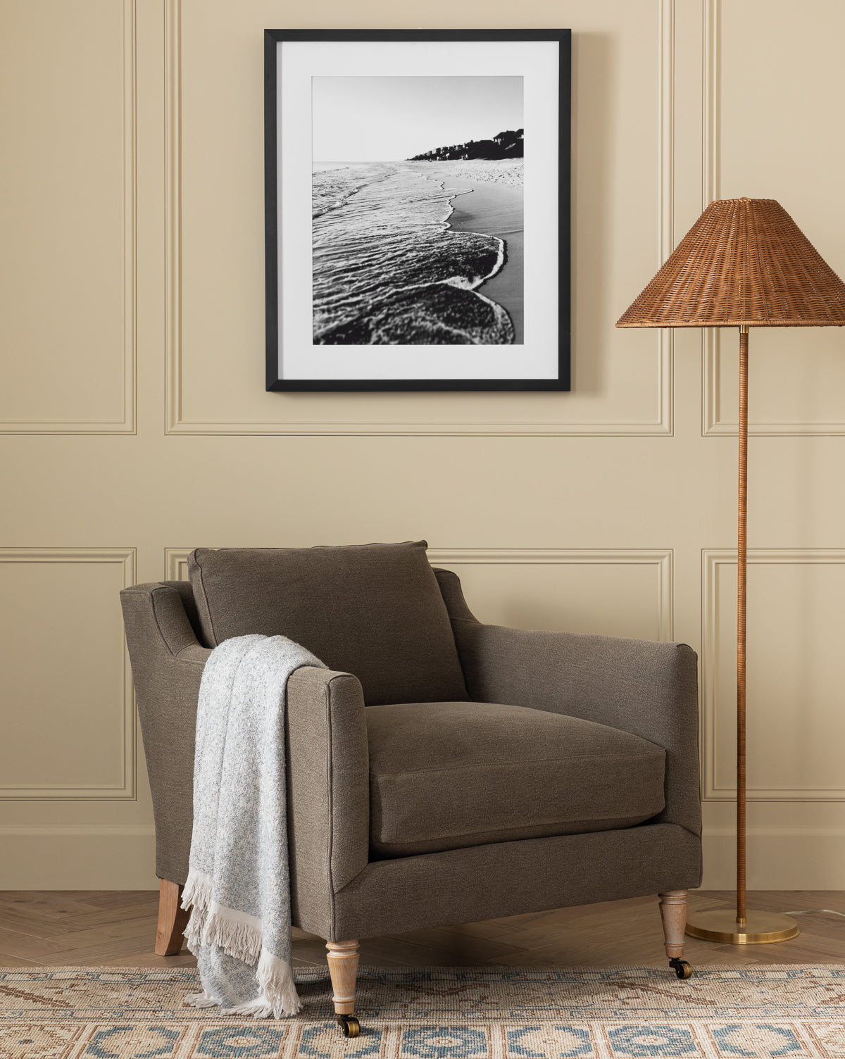 The Haverford Upholstered Lounge Chair by Rowe Fine Furniture, Inc. in brown, with a light gray throw, sits beside a wicker floor lamp. Above, a black-and-white beach photo hangs on beige paneling over a patterned rug. McGee & Co.
