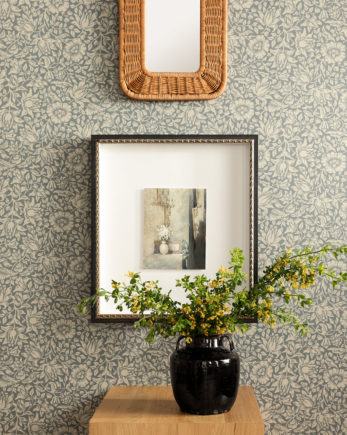 Certainly! Here’s your revised sentence with the exact string McGee & Co. blended at the end: A black vase with yellow flowers rests on a wooden table before the Morris & Co. x McGee & Co. Mallow Wallpaper by Sanderson Design Group, Inc.; above, a framed painting and rectangular wicker mirror add to the charming scene, all courtesy of McGee & Co.