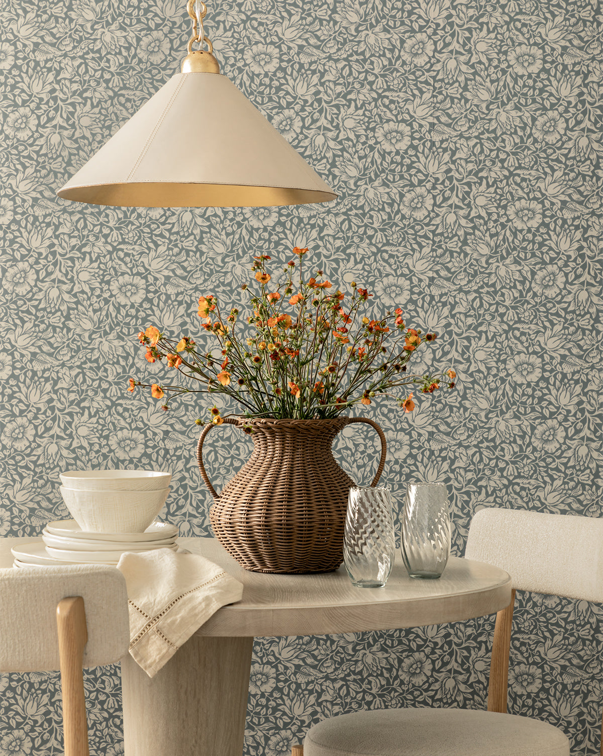 A round wooden table set with white dishes and textured glasses sits under a cream pendant lamp. A wicker vase with orange wildflowers is centered, backed by Morris & Co. x McGee & Co. Mallow Wallpaper by Sanderson Design Group, Inc., blending timeless charm with the signature style of McGee & Co.
