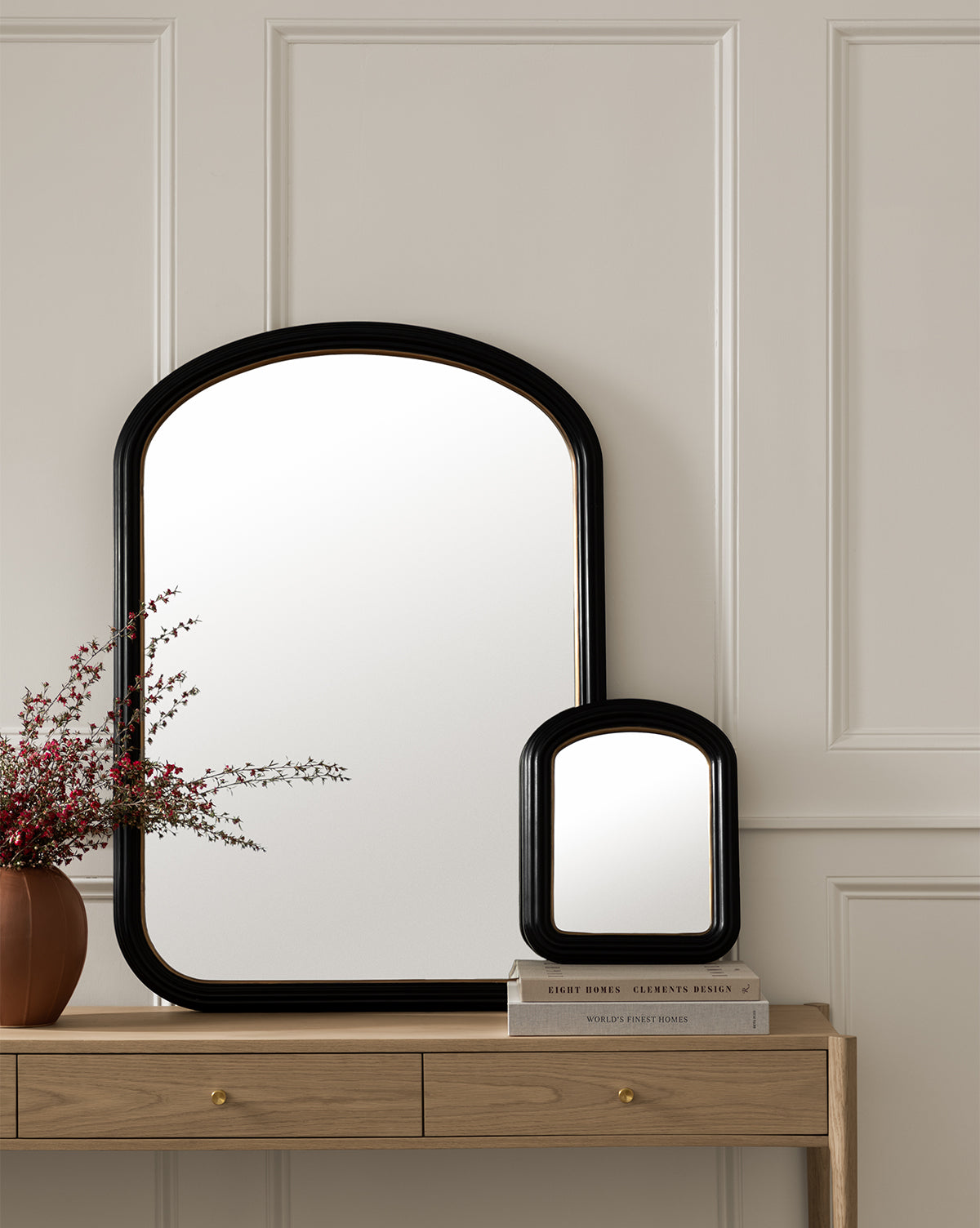 Two Samuel Wall Mirrors with black frames and arched tops hang over a wooden console table, which holds a brown vase of pink flowers and two stacked books, all set against a cream paneled wall, by McGee & Co.