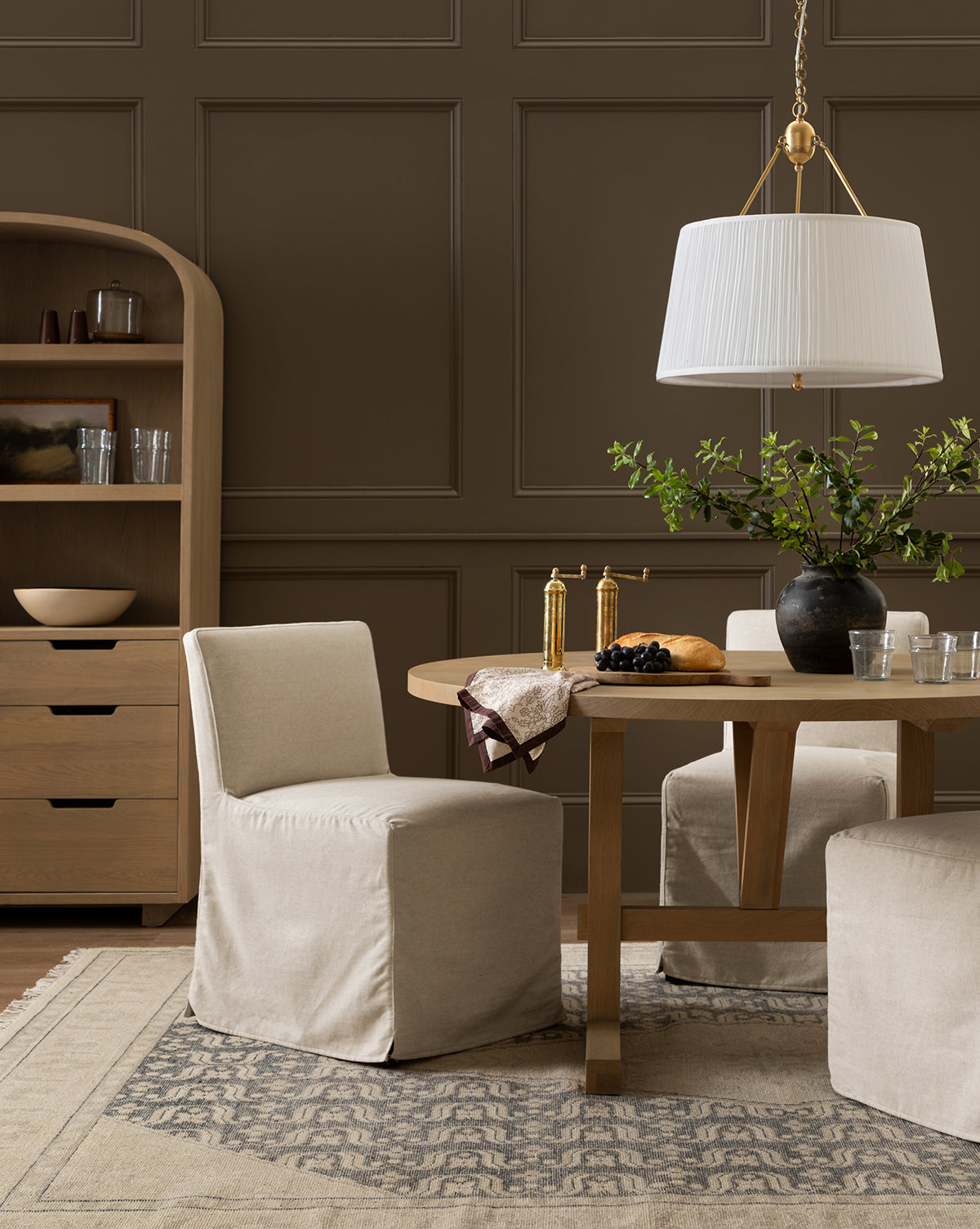 A modern dining room with neutral tones features a round wooden table, Olivier Slipcover Dining Chairs, a large pendant lamp, a patterned rug, a shelf sideboard, and a vase with green branches as the centerpiece, by McGee & Co.