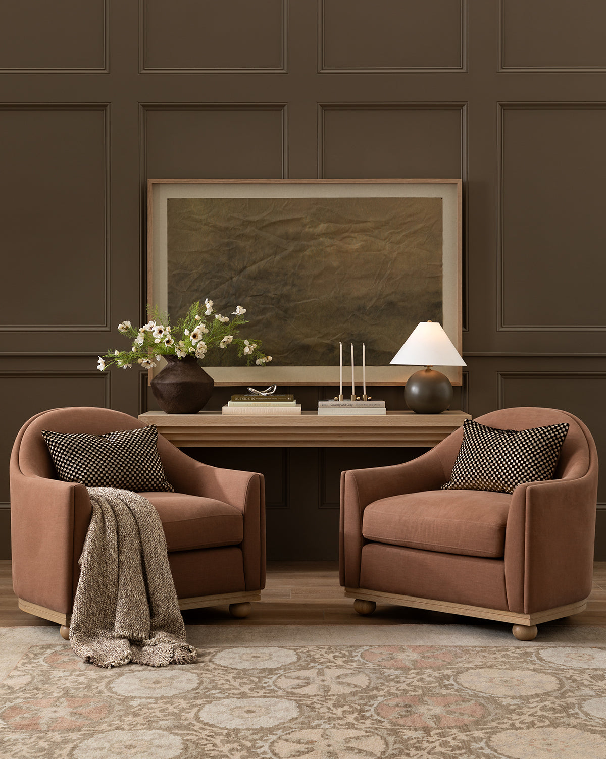 Two brown armchairs with patterned cushions and a textured throw sit atop a Suzani Hand-Knotted Wool Rug, facing a wooden console table adorned with flowers, candles, and a lamp, set against a paneled brown wall with large framed artwork, by McGee & Co.