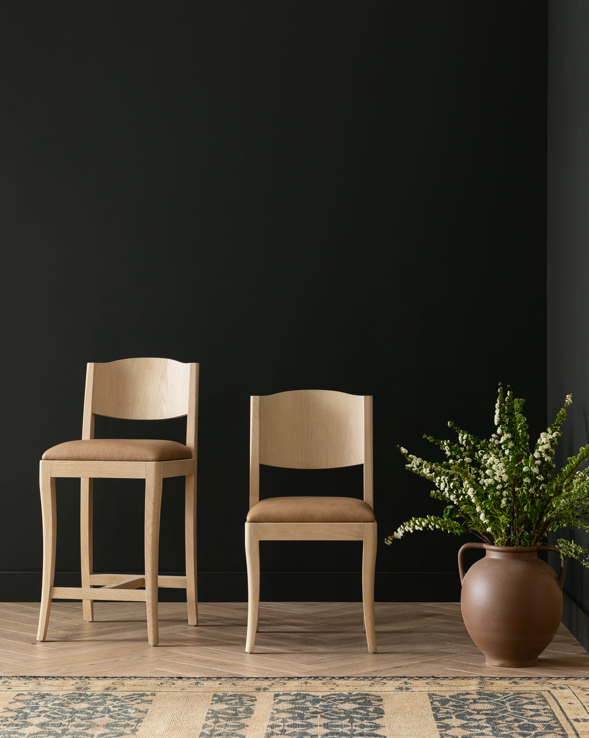 Two Shepherd Dining Chairs with white oak frames and tan upholstered seats are set on a wooden floor by a black wall. A large ceramic vase with green branches stands nearby, and a patterned rug appears in the foreground, by McGee & Co.