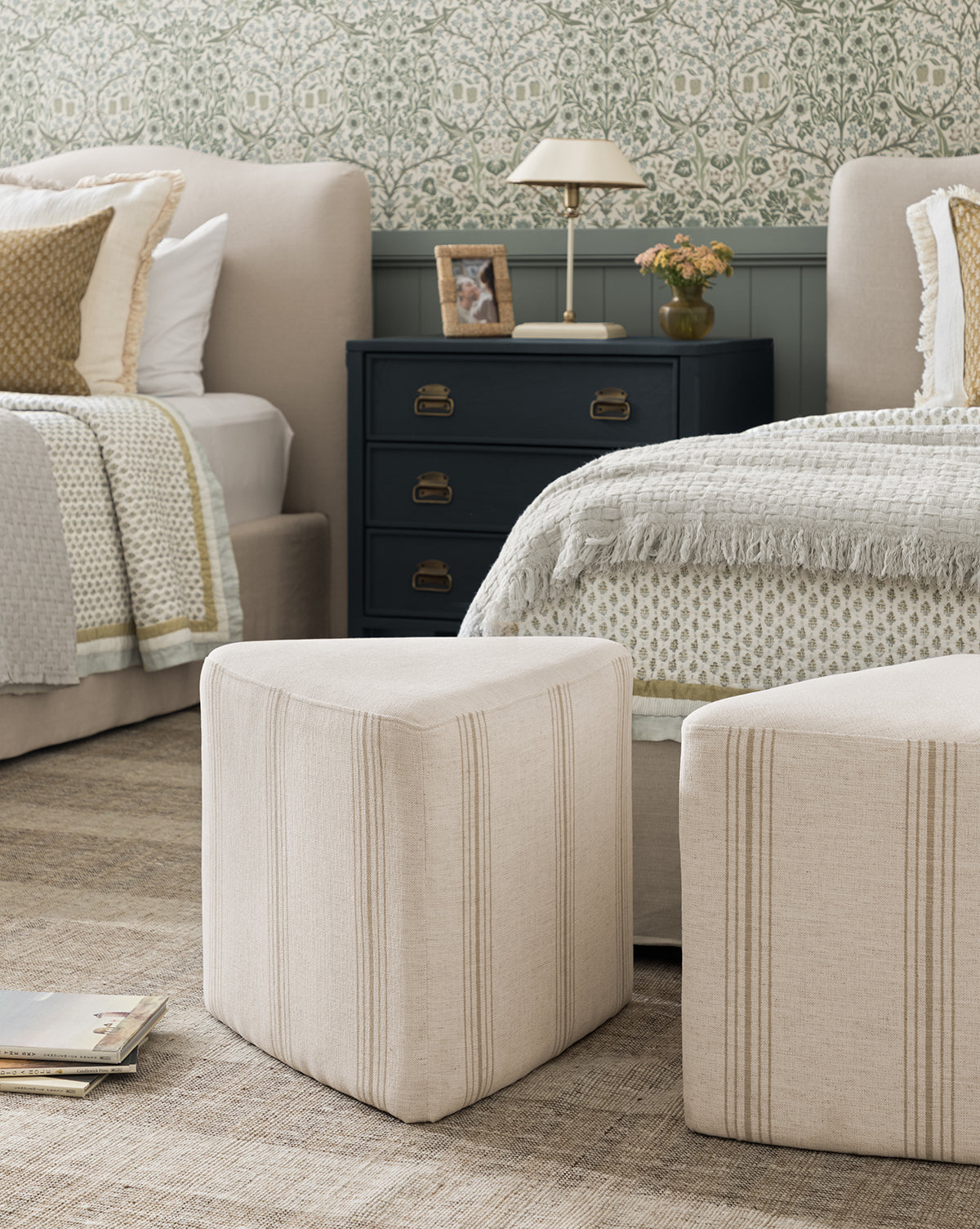 A cozy bedroom features two beige beds with textured bedding, a black nightstand with a lamp and flowers, two Jacques Ottomans by Dongguan City Baby Furniture Co., Ltd in crypton striped fabric, patterned wallpaper, and magazines on the woven rug—great for kids’ rooms, finished with accents from McGee & Co.