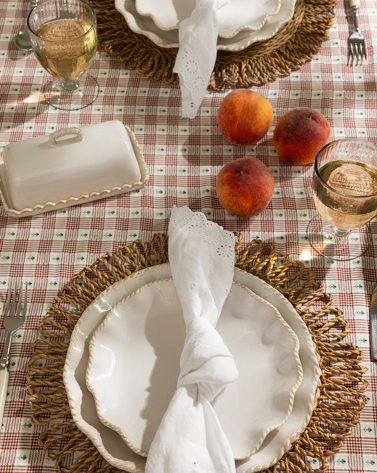 A table for two with scalloped white plates, woven placemats, wine glasses, a covered dish, three peaches, and McGee & Co. x Loeffler Randall Delilah Broderie Napkins (Set of 4) on a pink plaid Loeffler Randall tablecloth, by McGee & Co.